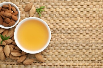 Almond oil in bowl, nuts and leaves on wicker table, flat lay. Space for text