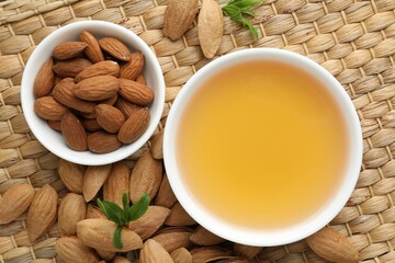 Almond oil in bowl, nuts and leaves on wicker table, flat lay