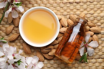 Almond oil in bowl, bottle, nuts, flowers and leaves on wicker table, flat lay