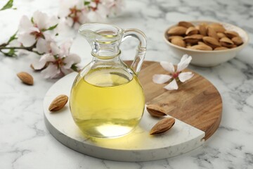 Almond oil in jug, nuts and flowers on white marble table, closeup
