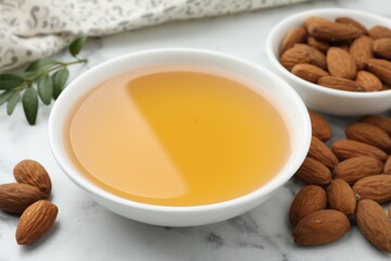Almond oil in bowl and nuts on white marble table, closeup