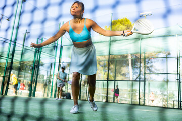 View through tennis net of focused young female padel player hitting volley to return ball to...