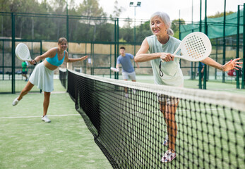 Senior woman in shorts playing padel tennis on court with young woman. Racket sport training...