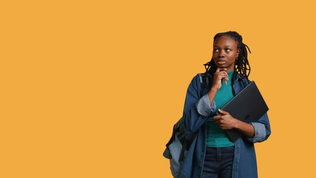 Pensive woman carrying laptop, doing thinking gesture, memorizing information for school exam, studio background. Student holding notebook, remembering before university assessment, camera B