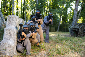 Paintball players of one team in camouflages and masks aiming with gun in shootout outdoors