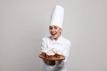 Happy professional confectioner in uniform holding delicious cupcakes on light grey background
