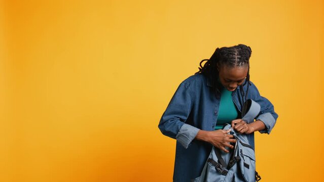 Portrait of smiling woman rummaging through backpack, looking for school notebook and notes, isolated over studio background. Cheerful girl taking out university supplies out of rucksack, camera B