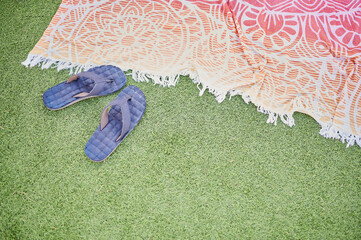 Pair of blue flip flops next to a colorful sarong spread on the grass of the pool to sunbathe. Poolside concept. - obrazy, fototapety, plakaty