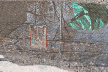 puddle reflecting a green tarp behind a chain link fence with faded no parking sign