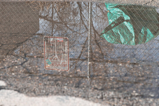 puddle reflecting a green tarp behind a chain link fence with faded no parking sign