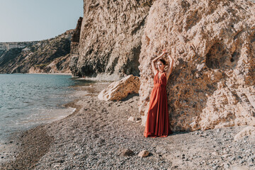 Woman red dress sea. Woman in a long red dress posing on a beach with rocks on sunny day. Girl on...