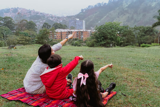International Children Day. Latina mother having fun with her young children outdoors. Sitting on the ground pointing at the landscape. Concept of family. Single mother with two small children.