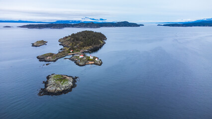 Island sitting in the ocean. A small collection of cabins and a lighthouse. In the distance two mountains meet at a small channel. 