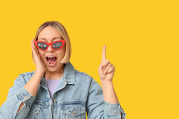 Surprised adult woman in sunglasses with words WOW pointing at something on yellow background