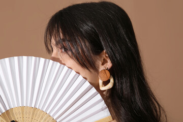 Young pretty woman with stylish earrings and hand fan on beige background