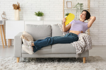 Fototapeta premium Young pretty woman with hand fan suffering from heat and lying on sofa in living room