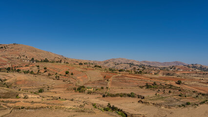 Rural landscape of Madagascar. Agricultural terraces of fields on red soil hillsides.  Single-storey residential buildings among green trees.Clear blue sky. Copy Space