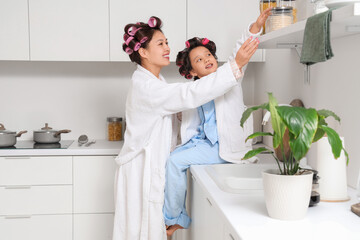 Little Asian girl and her mother with hair curlers in kitchen