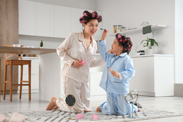 Little Asian girl and her mother with hair curlers applying mascara in kitchen