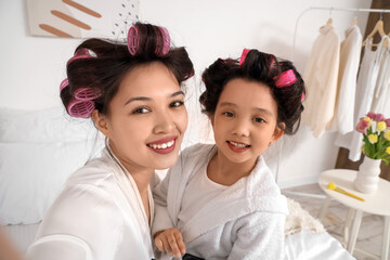 Cute little Asian girl and her mother with hair curlers taking selfie in bedroom, closeup