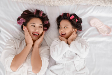 Cute little Asian girl and her mother with hair curlers lying on bed, top view