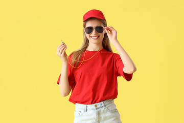 Young pretty female lifeguard in cap and sunglasses holding whistle on yellow background