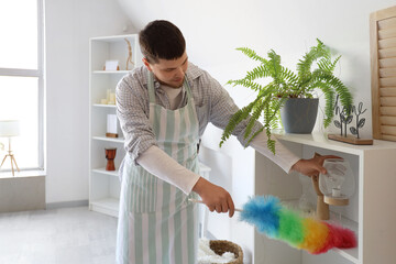 Young bearded man cleaning shelf with pp-duster at home