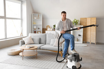 Young bearded man with vacuum cleaner having fun at home
