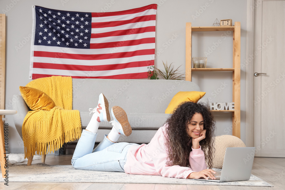 Wall mural Young African-American woman using laptop with USA flag on wall at home