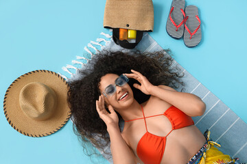 Young African-American woman with sunscreen cream and beach accessories lying on blue background,...
