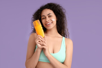 Young African-American woman with sunscreen cream on her face against lilac background