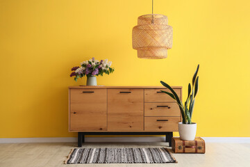 Vase with flowers on wooden cabinet, lamp and houseplant near yellow wall in room