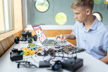 Schoolboy watching code on screen and using touch pad of laptop while learn technology in STEM class. On table put laptop, controller, electric wire, battery charger, and robotic vehicle. Edification.