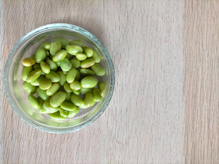 Cooked fresh soybeans (Glycine max) in a glass bowl on a light brown wooden surface, overhead shot