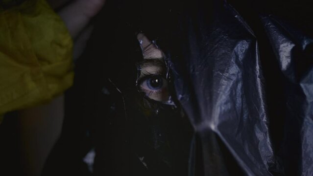 Portrait: the head of a girl in a black polythene bag. The face of the woman in the bag. The blue eye of a female through a hole in a plastic bag
