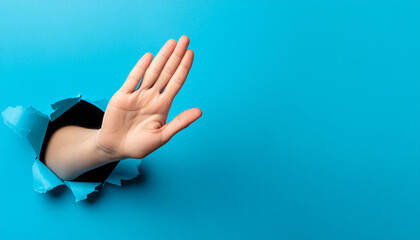 Hand making a stop sign, coming out of a torn blue paper hole. Concept of stop