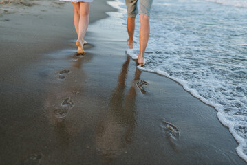 Down view of bare feet on sand beach. Couple in love walking barefoot on water with big waves on...