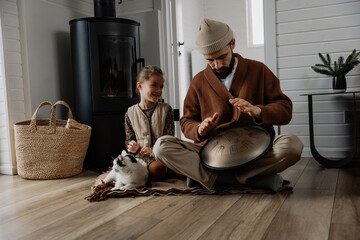 father and daughter spend time together sitting next to each other and playing handpan, share home time, playing with a cat and warming themselves by a hot fireplace