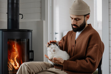 stylish bearded man with his home cat sitting near fireplace