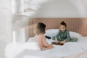 two little sisters aged 3 and 6, dressed in pajamas, are playing on the bed in the morning. Photo from the window