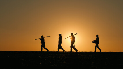 A family of farmers with equipment walks through the field. Silhouettes at sunset