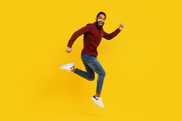 Fototapeta na wymiar Indian man wearing a red sweater is captured mid-jump in the air. His arms are raised, and his legs are bent as he springs upwards. The background is yellow, emphasizing the mans movement.