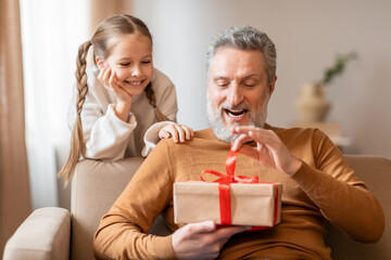 Senior man and a young girl are seated on a couch, both looking excitedly at a wrapped present...
