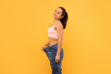 A cheerful young woman stands against yellow backdrop, holding her overly large jeans to show her successful weight loss journey. Her bright smile and confident pose convey a sense of achievement.