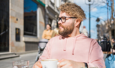 Millennial bearded man having breakfast at table of street cafe on spring day, drinking warm...