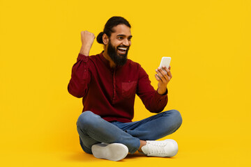 Indian man is seated on the floor, holding a cell phone in his hands. He appears focused on the...