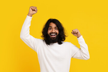 A bearded young Indian man with long hair exudes excitement and joy as he raises his fists in a...
