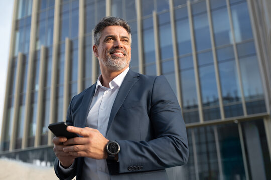 Portrait of smiling executive ceo manager. Latin adult man in suit using smartphone application app device for business at office outdoors. Successful mid age senior businessman holding mobile phone. 