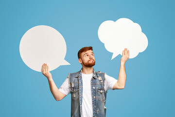 Young redhaired man having complex choice holding two speech bubbles on blue background, mockup,...