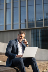 Mature businessman working remotely on laptop, sitting outdoors, talking on mobile phone with...
